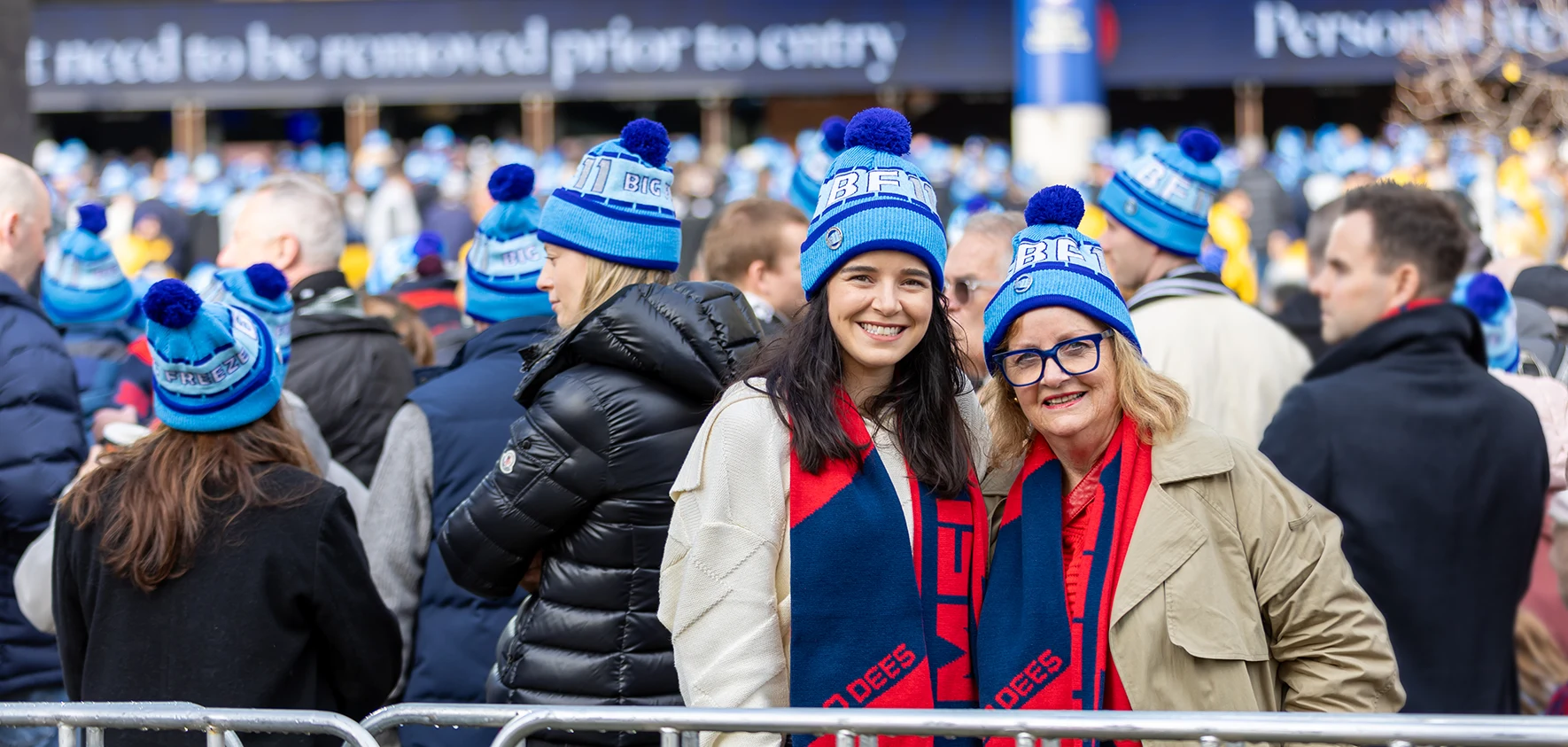 Two women in Big Freeze beanies and Melbourne Demons scarves smile together in in MCG Gate 2 crowd